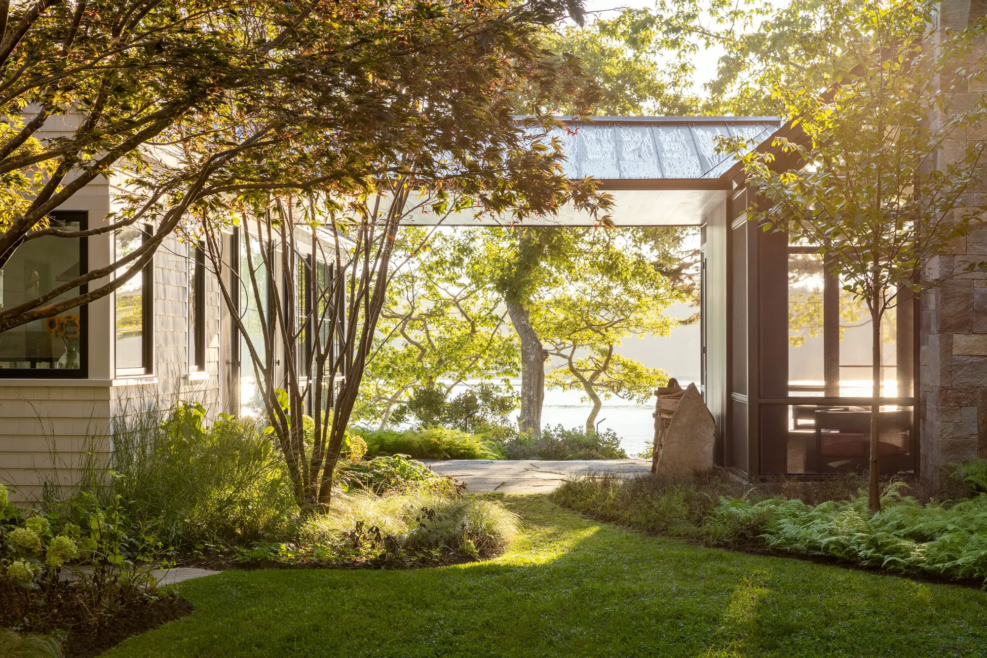 Sunlit garden with trees and grass between two modern buildings overlooking a lake.