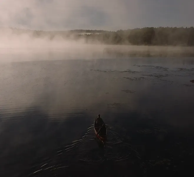 Two people paddling a canoe on a misty lake at dawn or dusk with forest in the background.