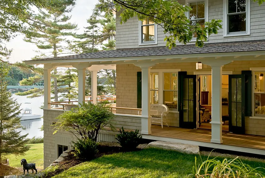 Porch of a house with white columns, wicker chair, green lawn, and trees by a lake with docked boats.