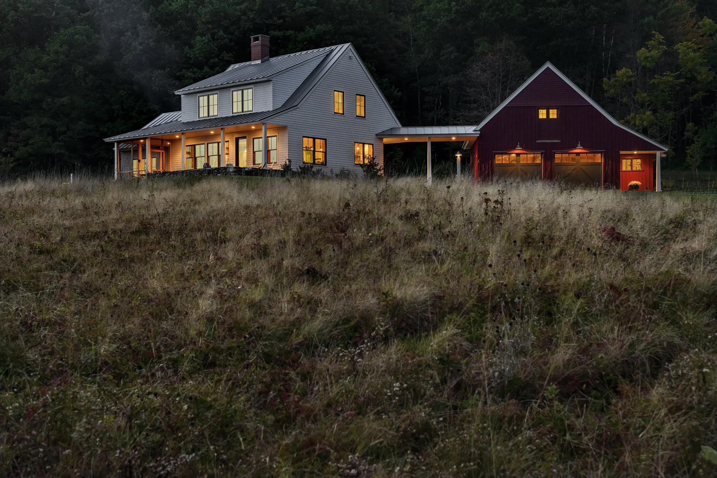 White farmhouse with lit windows next to red barn in a field at dusk with forest background.