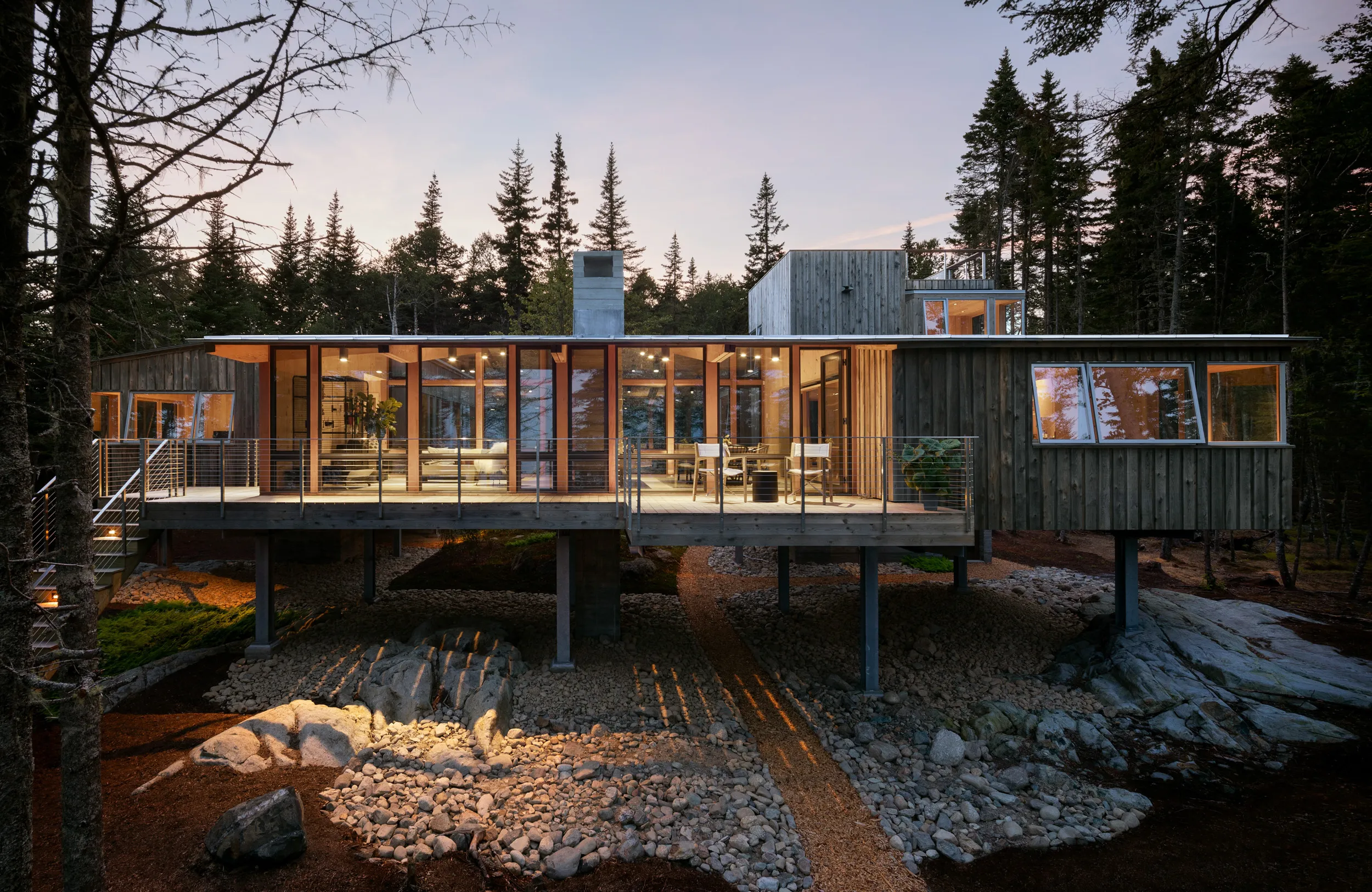 Modern wooden house on stilts with large windows, surrounded by forest at dusk.