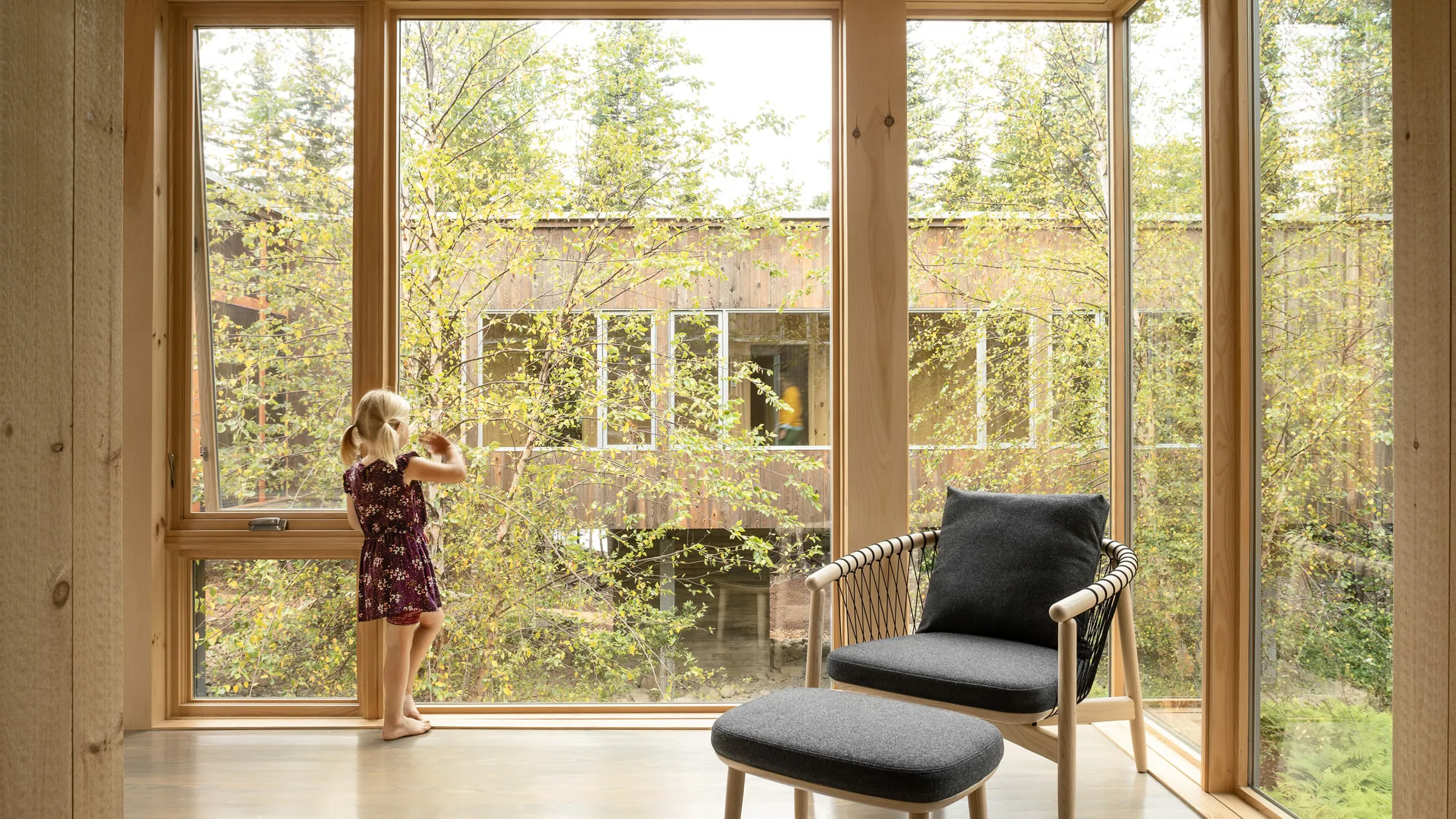 A girl in a purple dress standing and looking out a large window at green trees outside.