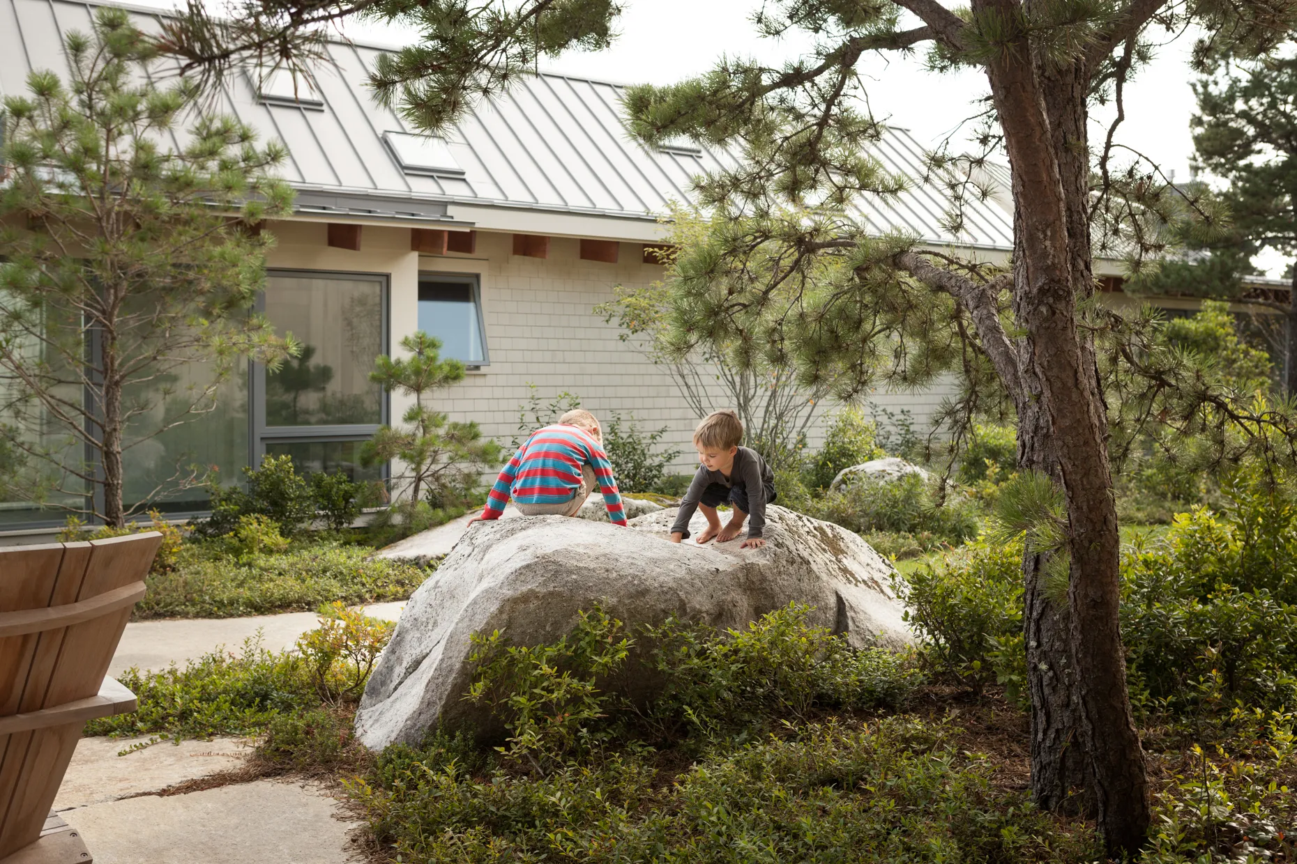 Two young children playing and climbing on a large rock in a garden outside a modern house.