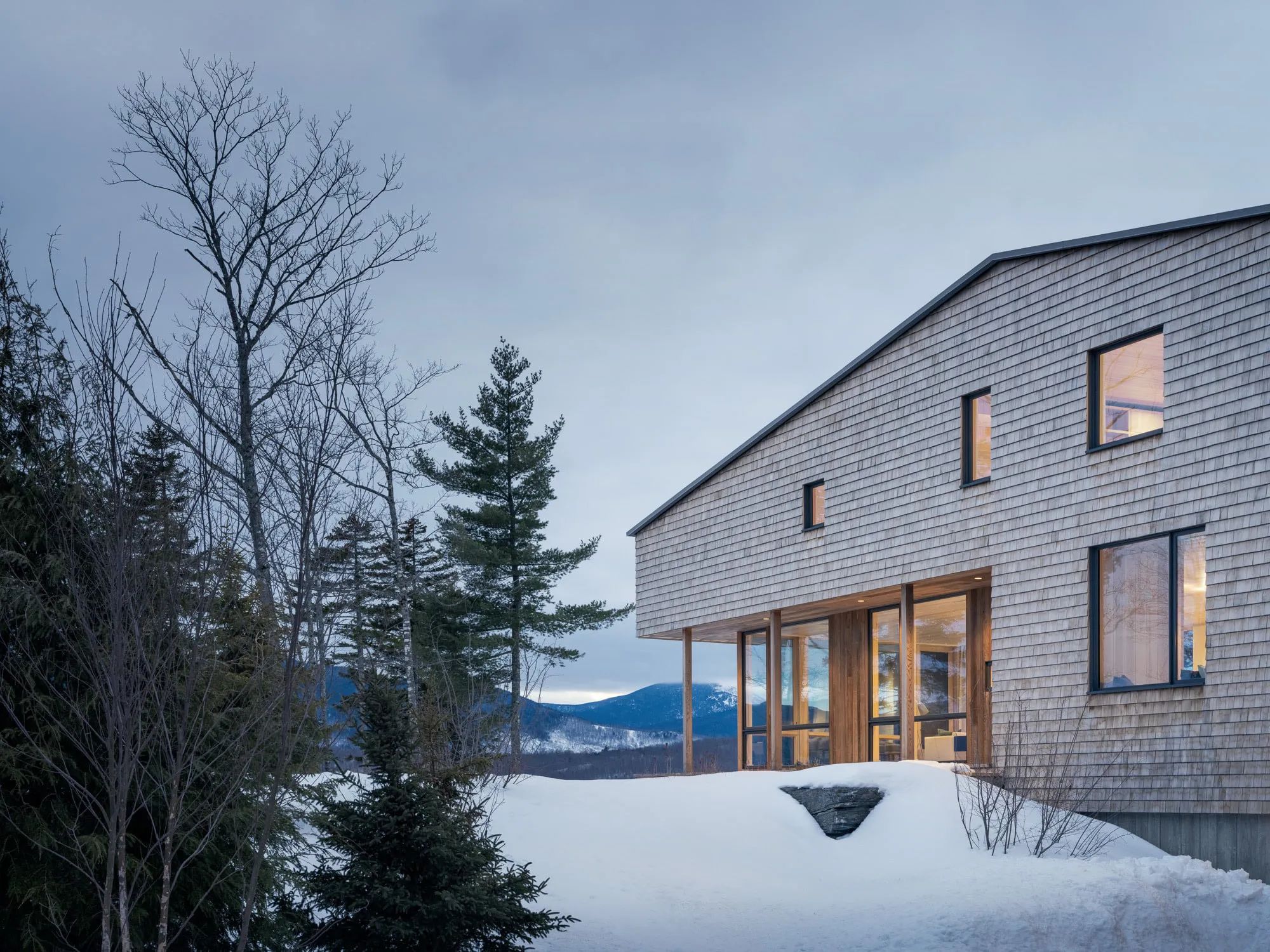Modern house with wooden shingles by snow-covered ground and trees under a cloudy sky.