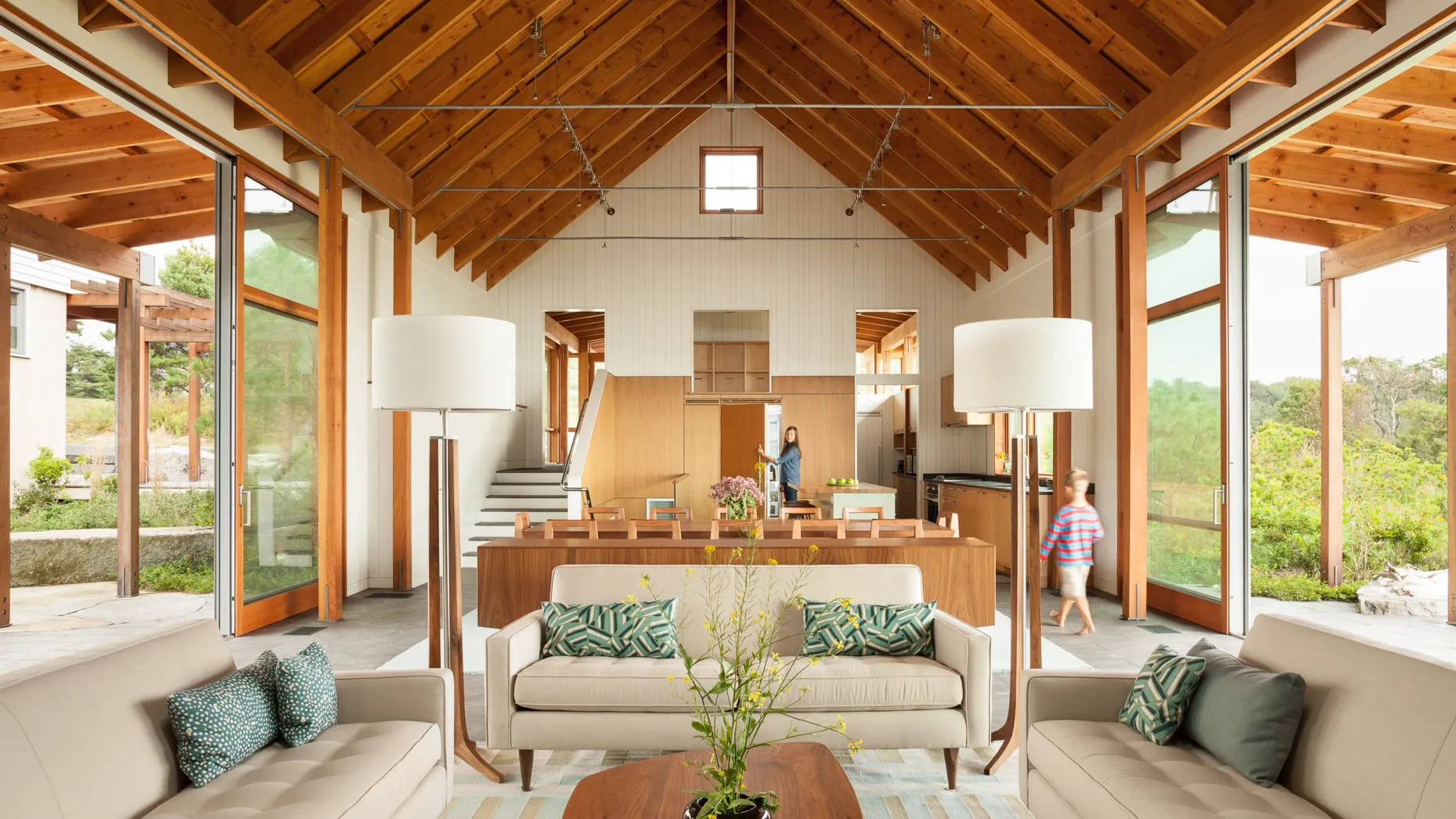 Open living room with beige sofas, wooden beams, large windows, and two people near the kitchen area.