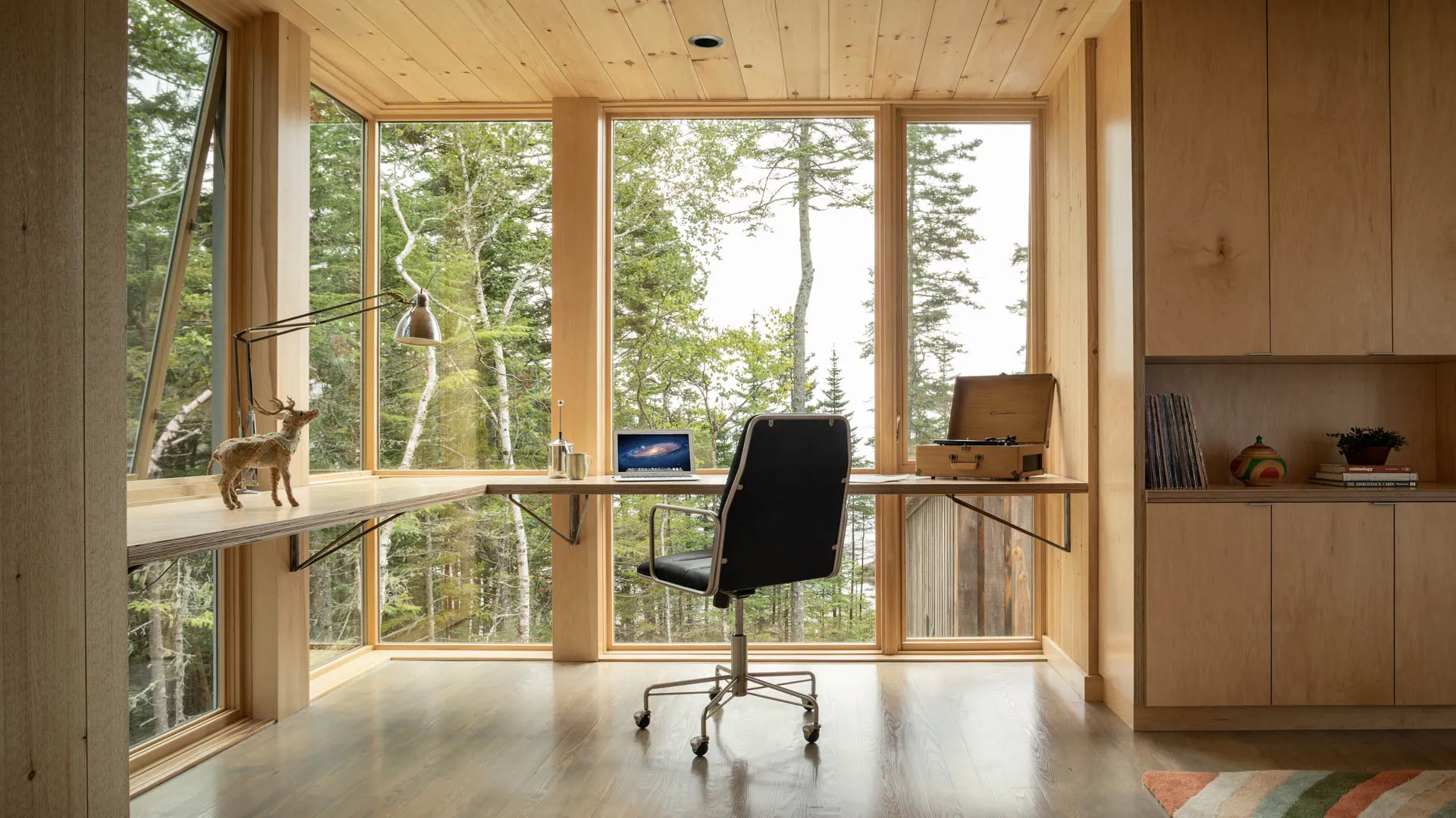 Minimalist wooden home office with black chair, laptop, record player, and large windows showing forest.