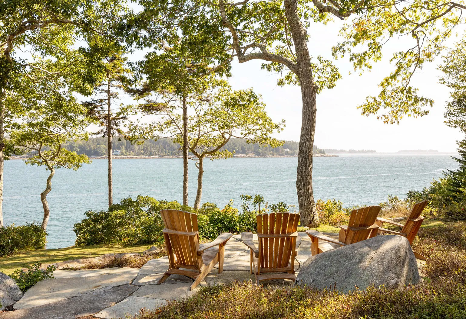 Four wooden Adirondack chairs on stone patio overlooking calm water surrounded by trees and shrubs.