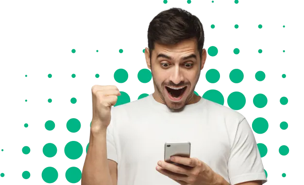 Hombre con camiseta blanca sonriente, mirando su teléfono móvil con emoción y puño levantado en señal de alegría.