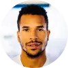 Hombre joven con barba y cabello rizado, sonriendo, en un fondo desenfocado.