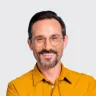 Hombre con barba y gafas, sonriente, lleva una camisa amarilla sobre fondo claro.