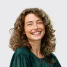 Mujer con cabello rizado y suelto, sonriente, vestida con blusa verde, fondo claro.