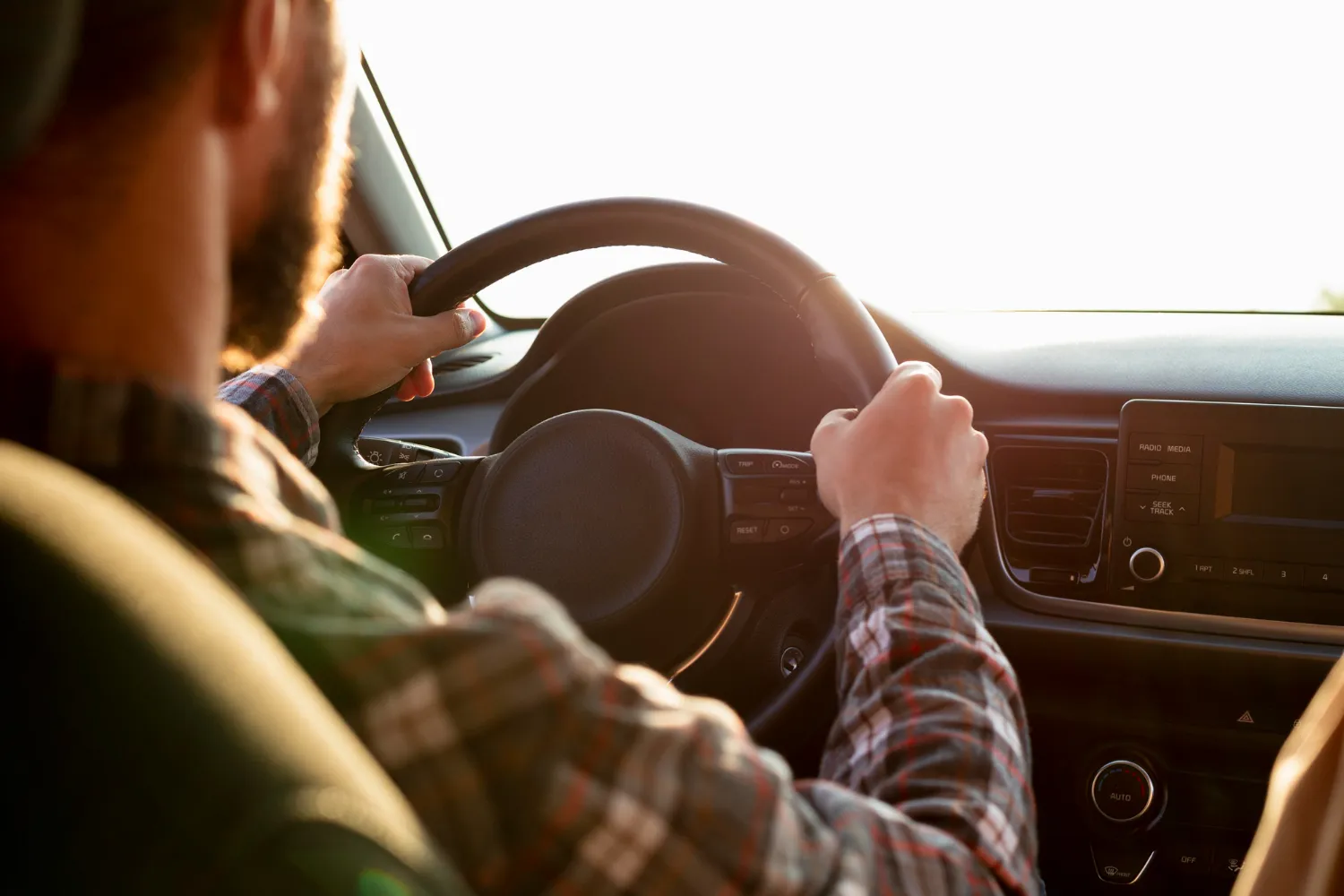 Hombre con camisa de cuadros sostiene el volante de un coche mientras conduce, con luz del atardecer iluminando el interior.