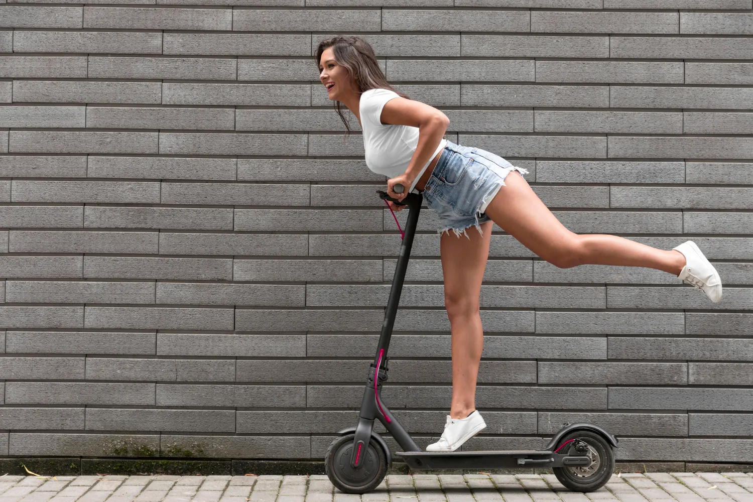 Mujer sonriente montando un patinete, con una pierna levantada y usando un short de mezclilla y camiseta blanca. Fondo de ladrillos grises.