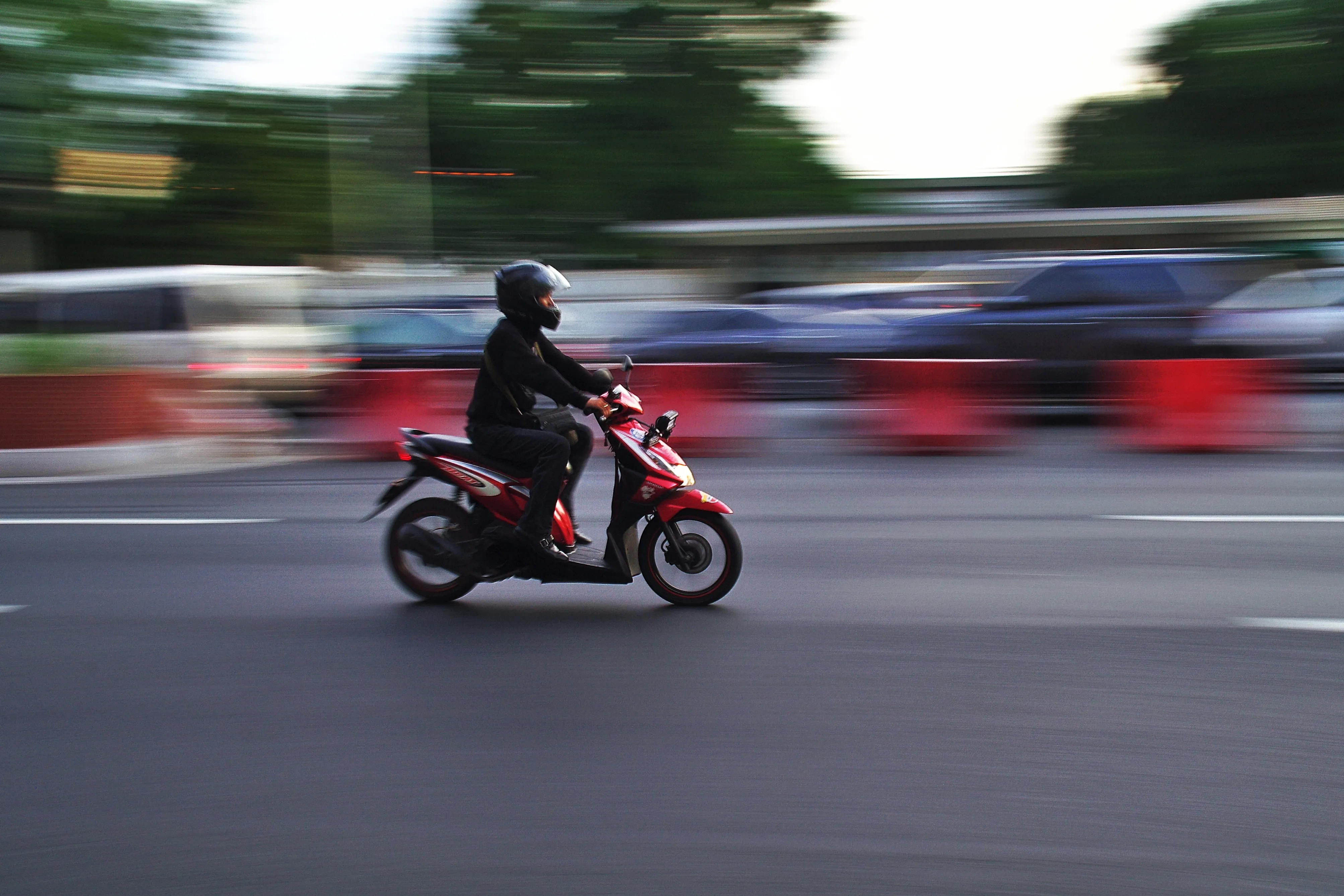 Motocicleta roja en movimiento, con un conductor vestido de negro y casco, en una carretera con vehículos y barreras de seguridad.