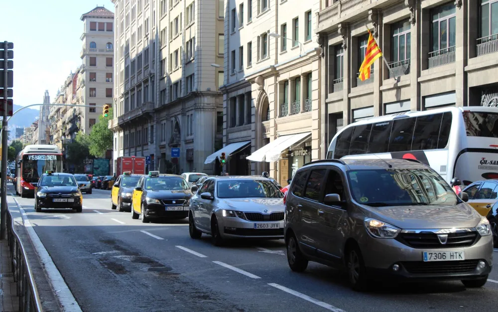 Tráfico en una calle de ciudad con coches, taxis y un autobús, edificios altos y una bandera ondeando.