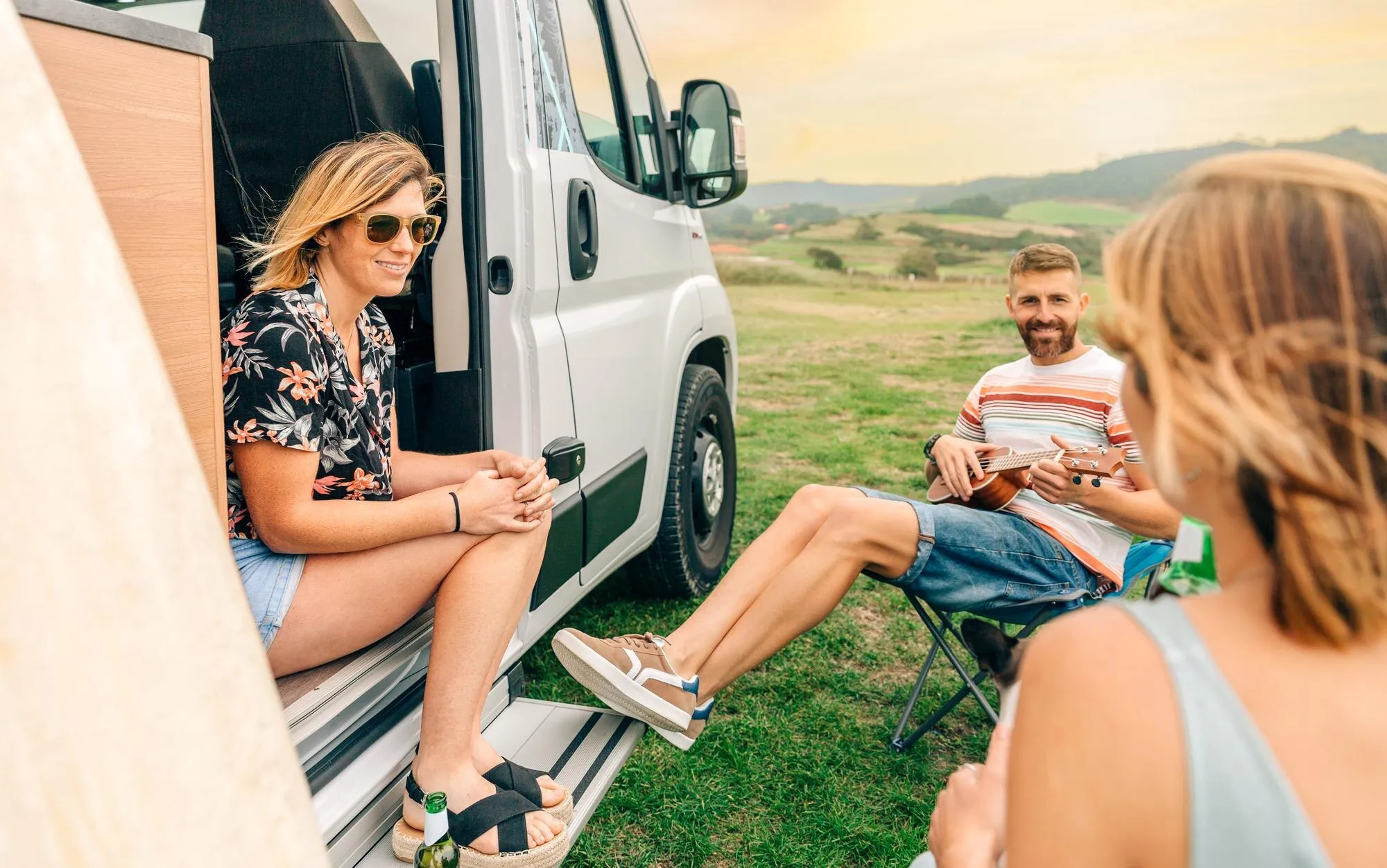 Pareja sentada junto a una autocaravana, disfrutando de un momento al aire libre. Hombre toca el ukelele mientras conversan.