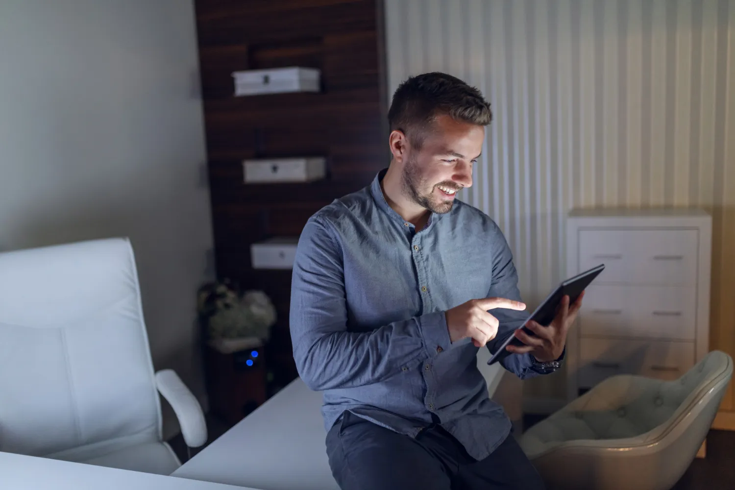 Hombre sonriente usando una tablet en un ambiente iluminado suavemente, sentado en una mesa con sillas blancas.