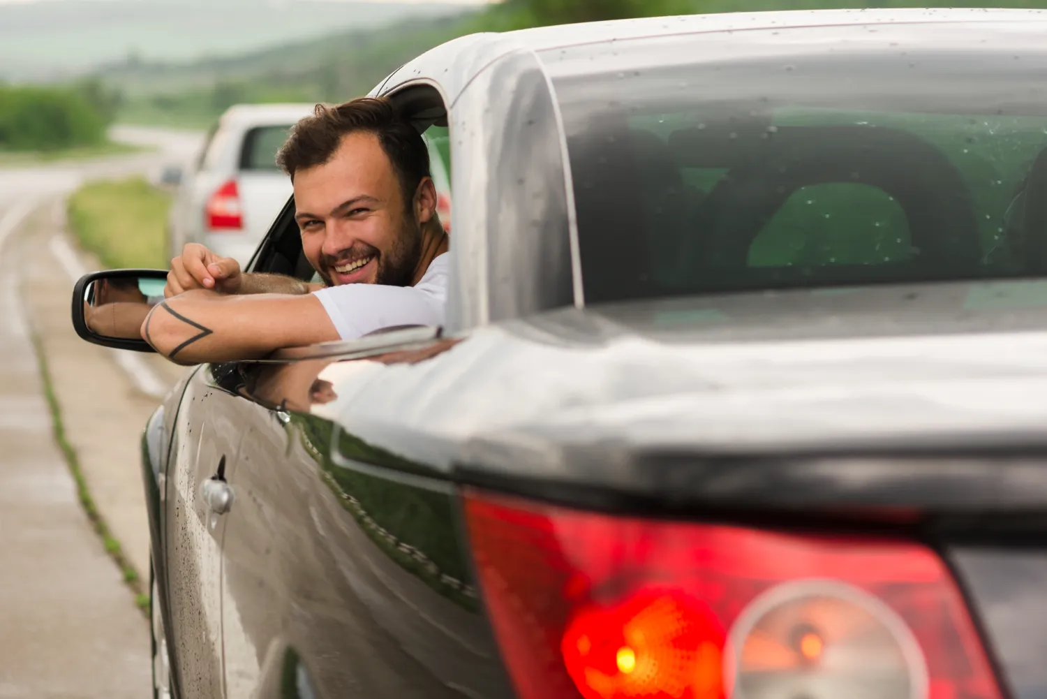 Hombre sonriente asomado por la ventana de un coche negro, con un paisaje verde y carretera de fondo.