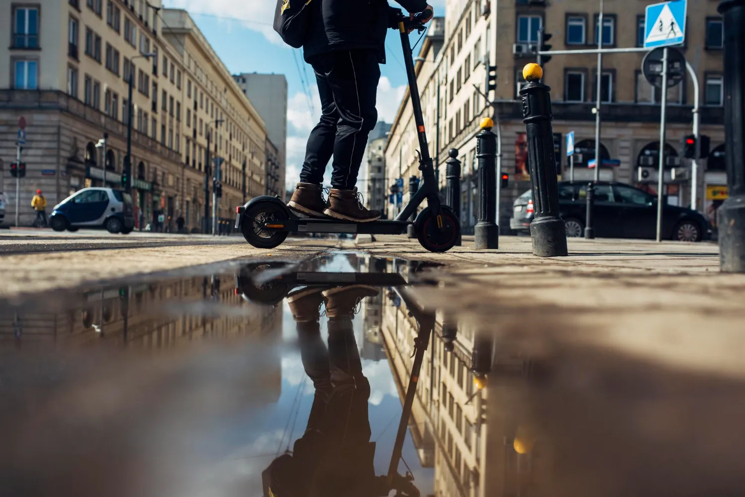 Persona sobre un patinete eléctrico, reflejada en un charco, con edificios y tráfico urbano de fondo.