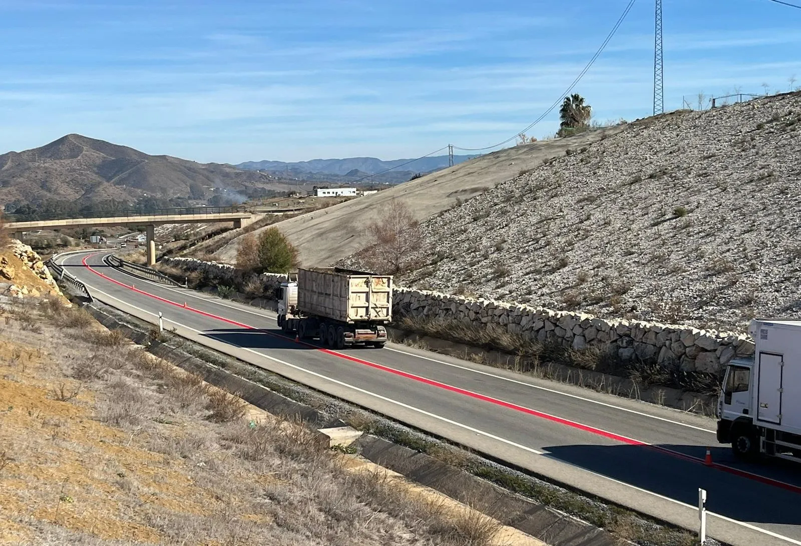 Camión circulando por carretera con líneas rojas en el asfalto, rodeado de montañas y vegetación escasa.