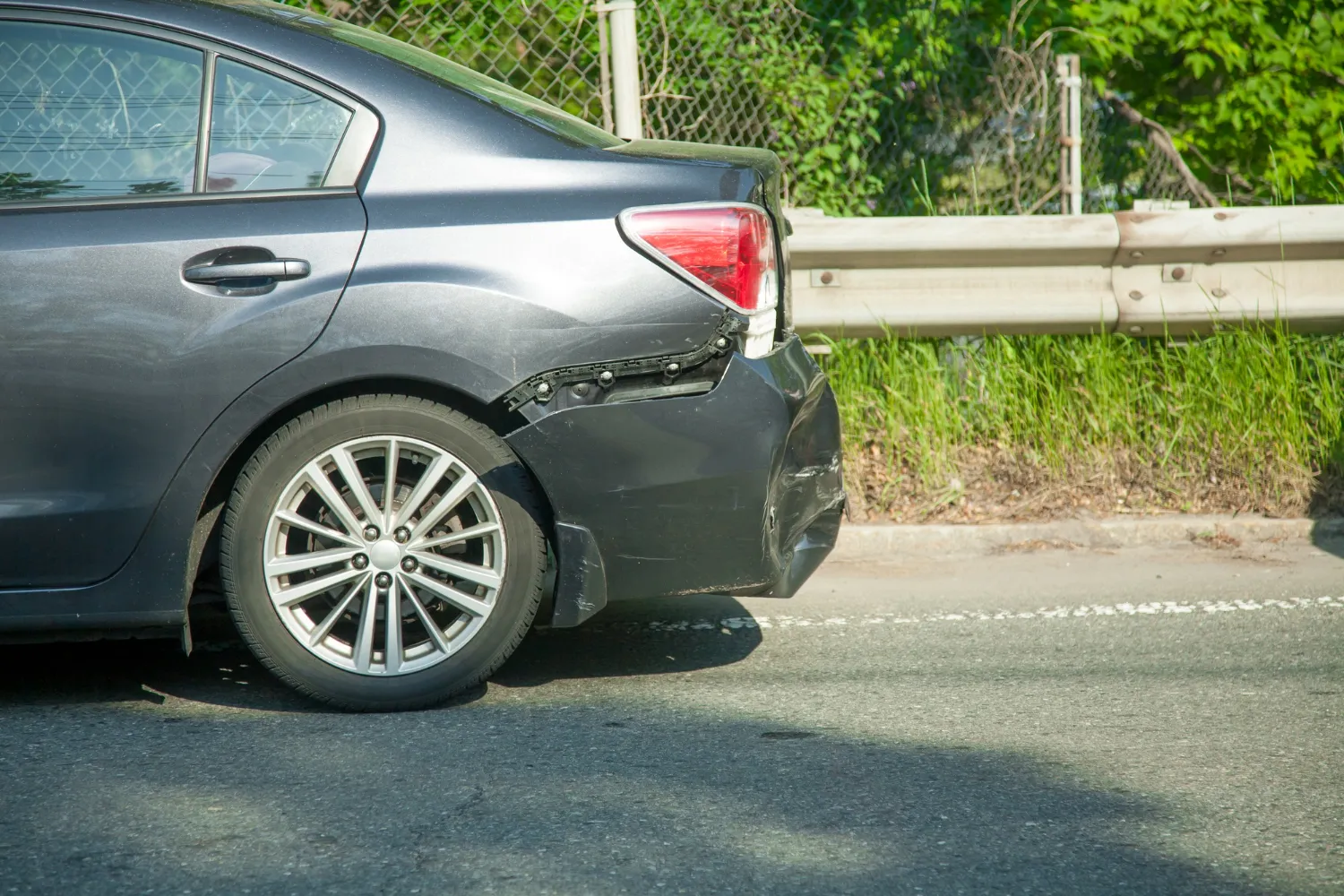 Auto gris con daños visibles en la parte trasera, cerca de una barrera de seguridad y vegetación al fondo.