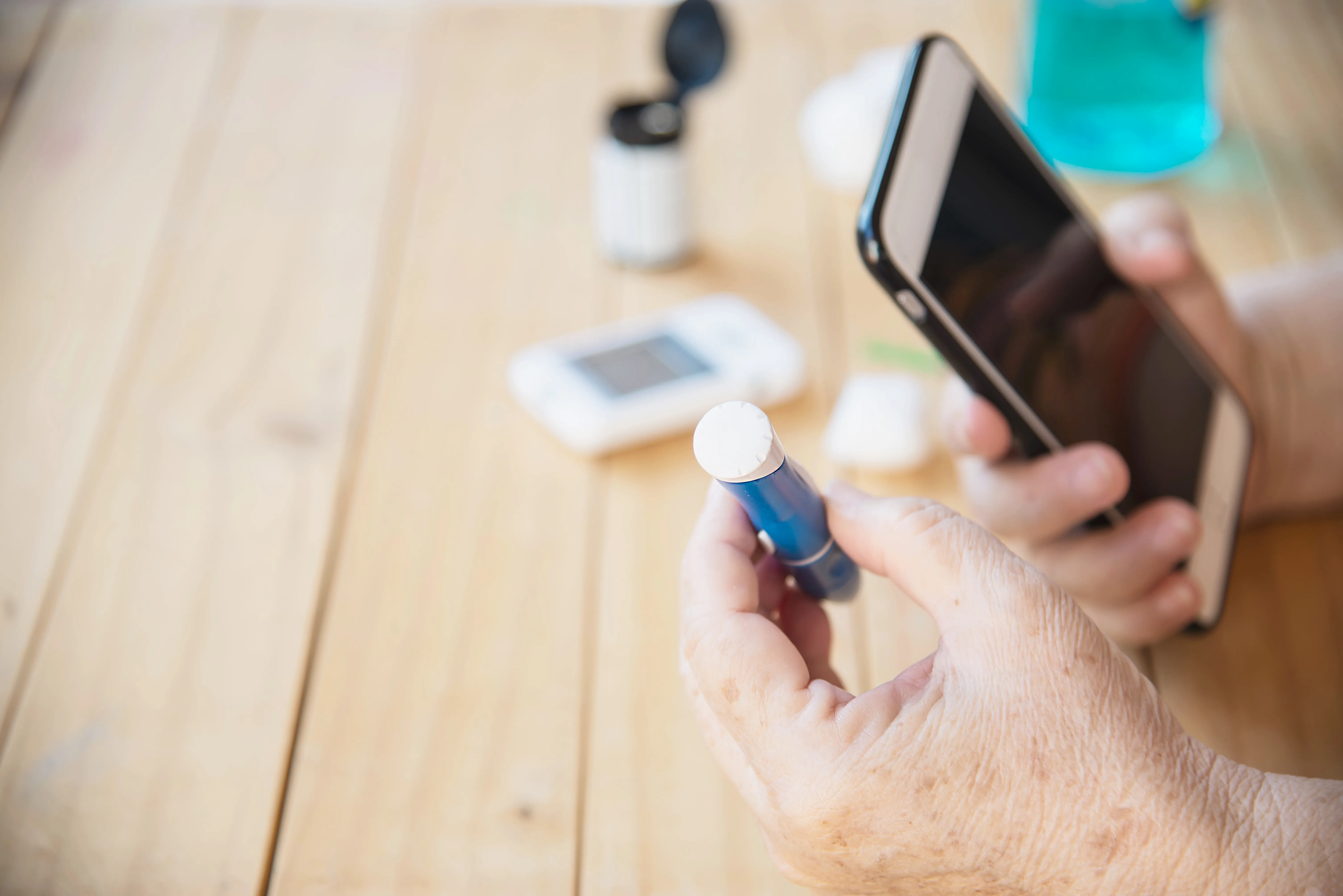 Mano sosteniendo un dispositivo de medición de glucosa, mientras se utiliza un teléfono móvil. Fondo de mesa de madera.