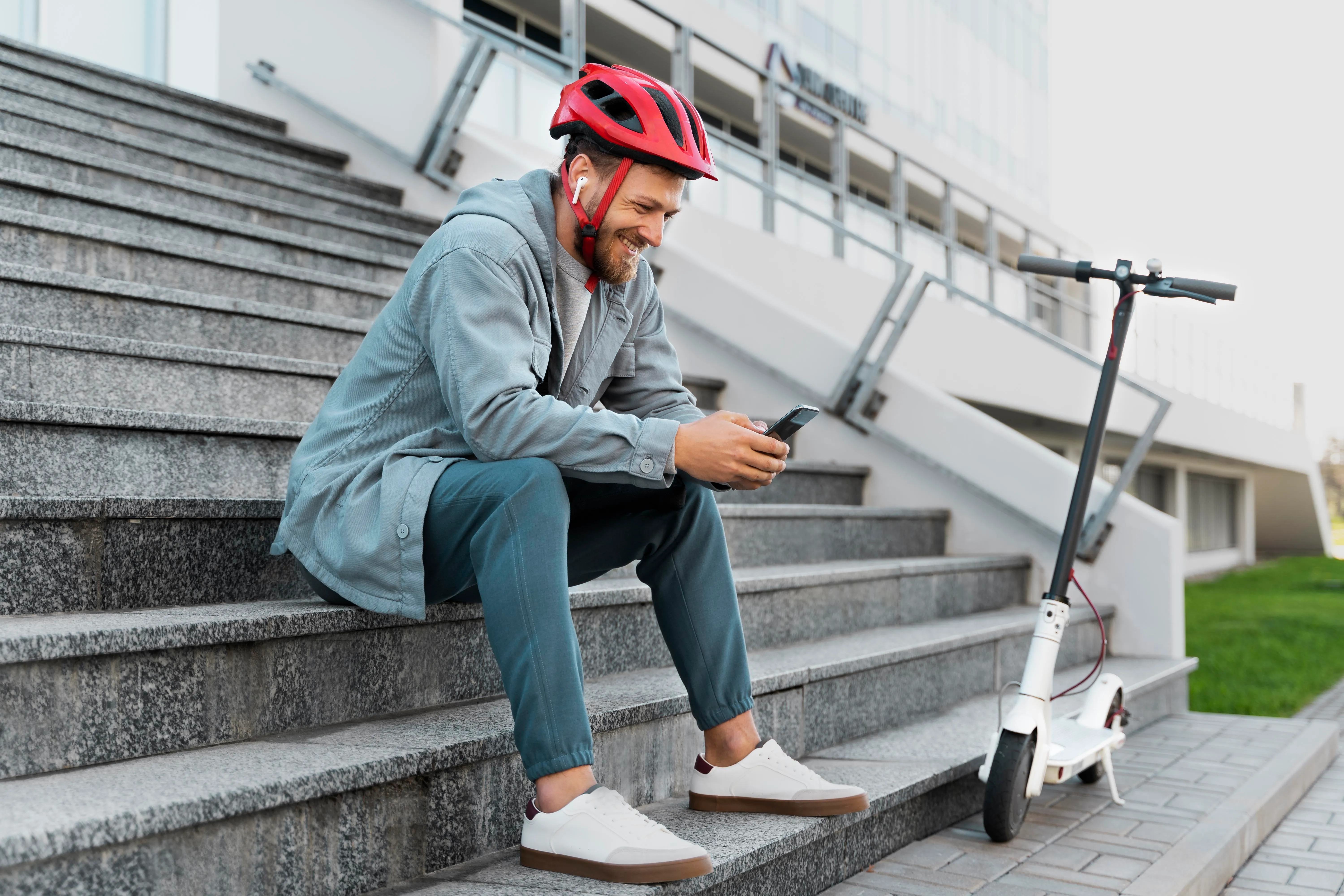 Hombre sentado en escaleras, usando un teléfono móvil, con casco rojo y patinete eléctrico a su lado.