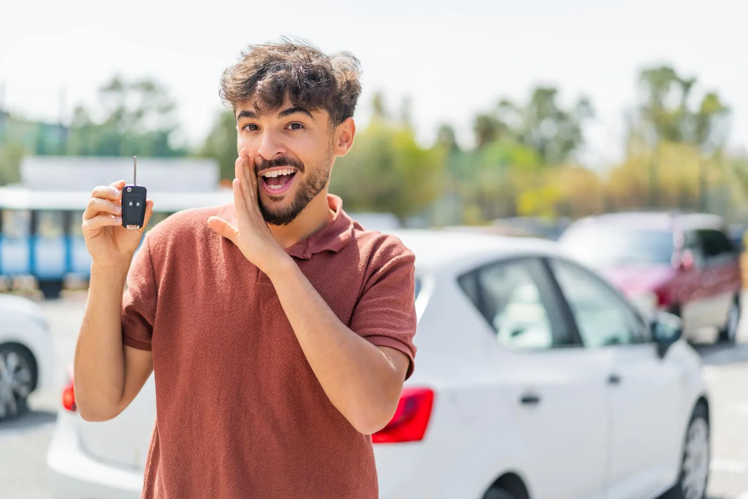 Hombre sonriente sostiene una llave de coche, con un fondo de vehículos estacionados y un ambiente soleado.