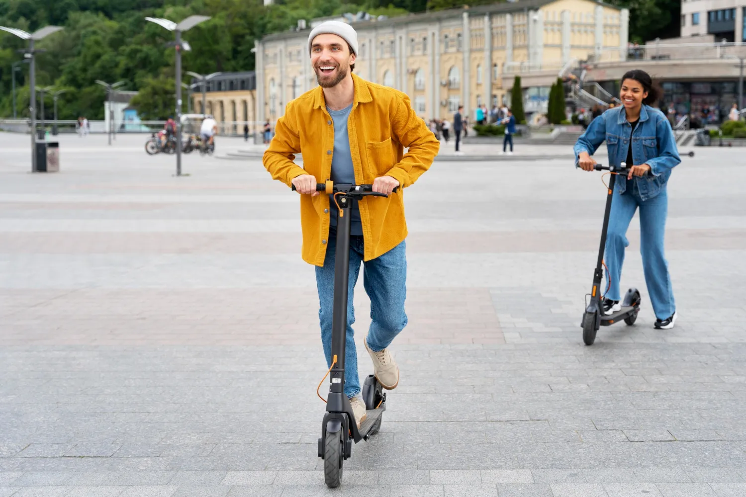 Hombre sonriente montando un patinete eléctrico, mientras una mujer lo sigue en un entorno urbano con pavimento y edificios al fondo.