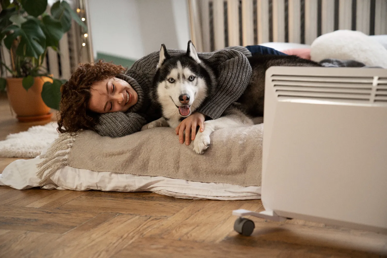 Mujer sonriente abrazando a un husky en una cama, junto a un calefactor, en un ambiente acogedor.
