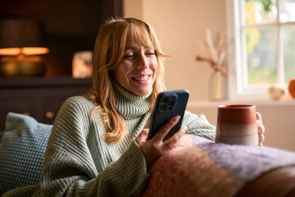 Mujer sonriente con suéter de cuello alto, sosteniendo un teléfono y una taza, sentada en un sofá acogedor.