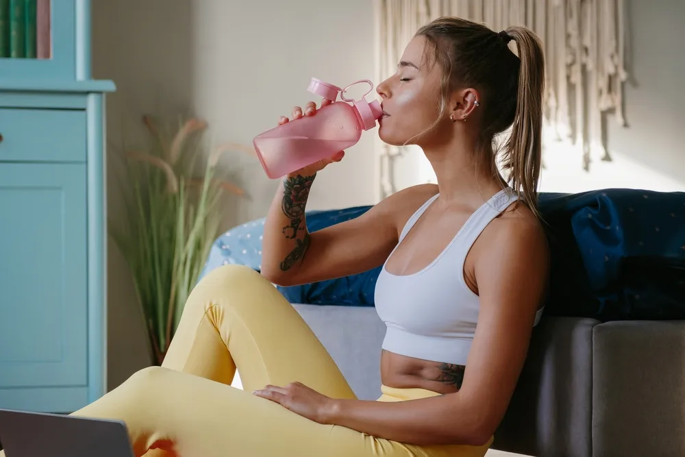 Mujer con top blanco y leggings amarillos bebiendo de una botella rosa, sentada en un sofá con un portátil frente a ella.