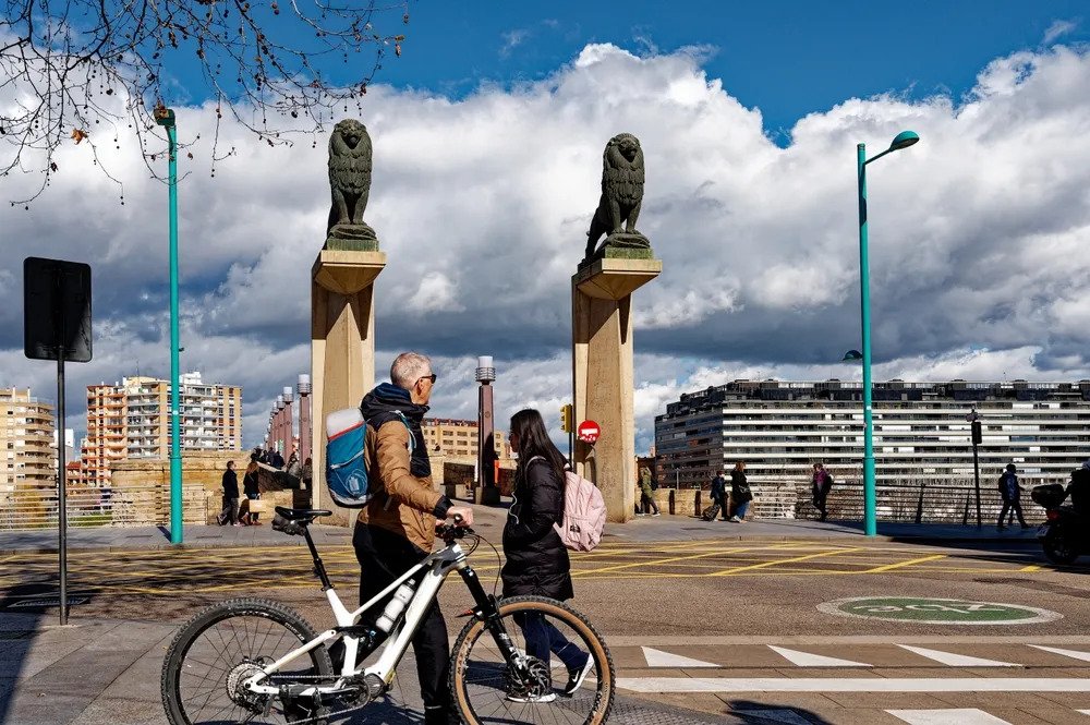 Pareja de personas en un puente, una con bicicleta, rodeados de edificios y nubes en el cielo.