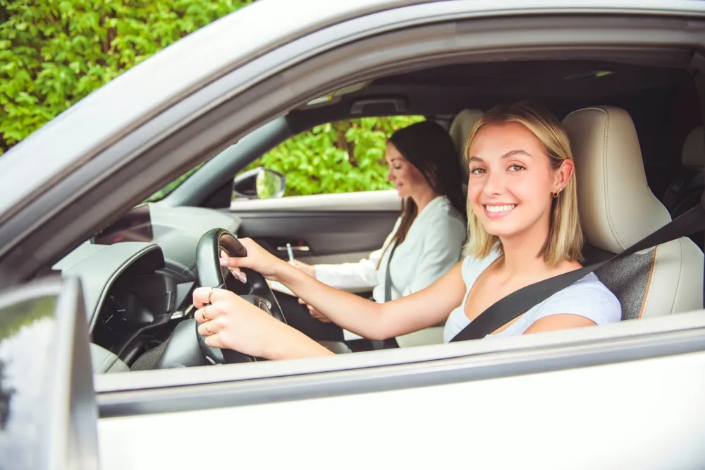 Joven sonriente al volante de un coche, acompañada por otra mujer en el asiento del pasajero. Fondo verde.