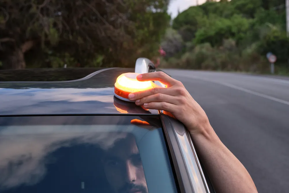 Una mano coloca una baliza V16 naranja sobre el techo de un coche, con un paisaje natural y carretera al fondo.