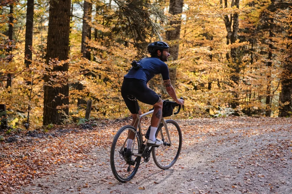 Ciclista pedaleando por un sendero de grava rodeado de árboles con hojas amarillas en un ambiente otoñal.