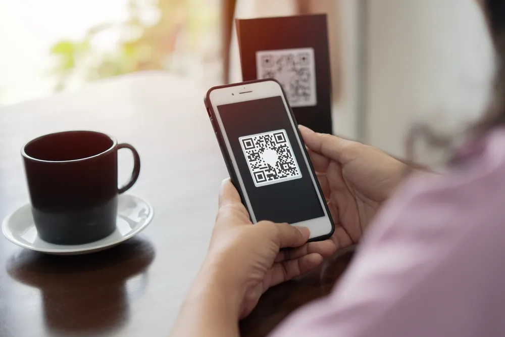 Persona sosteniendo un teléfono móvil que escanea un código QR, con una taza de café y un fondo desenfocado.