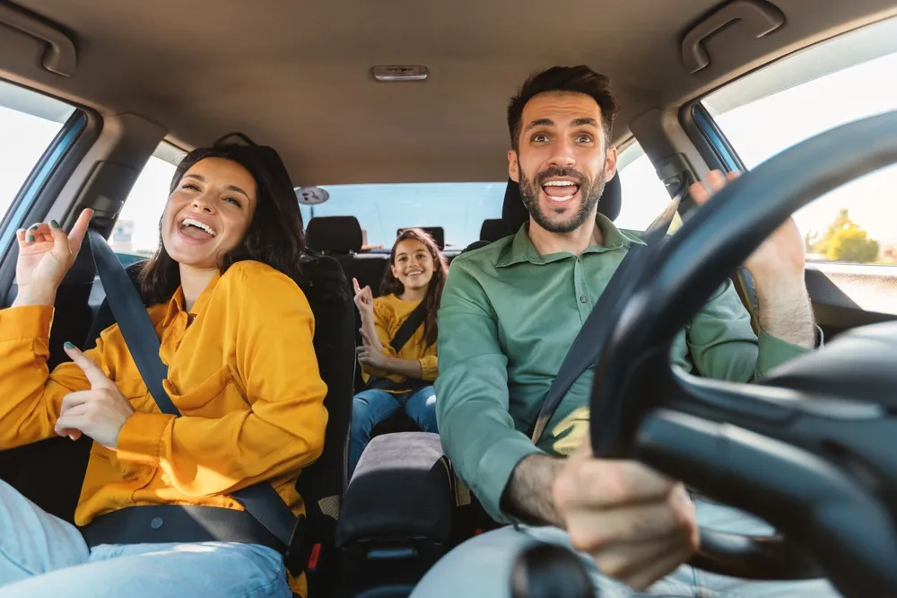 Tres personas sonrientes en un coche, disfrutando y bailando mientras conducen. La mujer a la izquierda lleva una camisa amarilla.