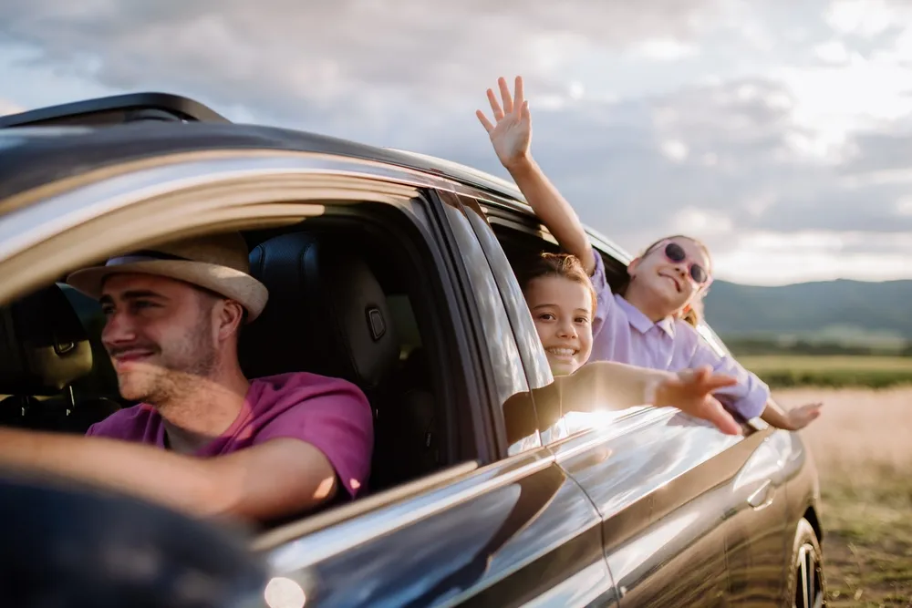 Hombre sonriente conduce un coche, mientras dos niños asoman por la ventana, disfrutando del paisaje y el aire libre.