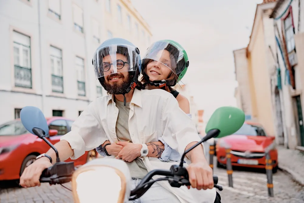 Pareja sonriente en una moto, ambos con cascos, en una calle con coches estacionados y edificios de fondo.