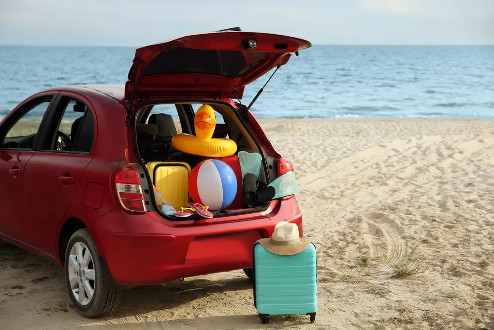Maletero de un coche rojo abierto, con maletas, una pelota de playa y un inflable de pato, frente a la playa y el mar.