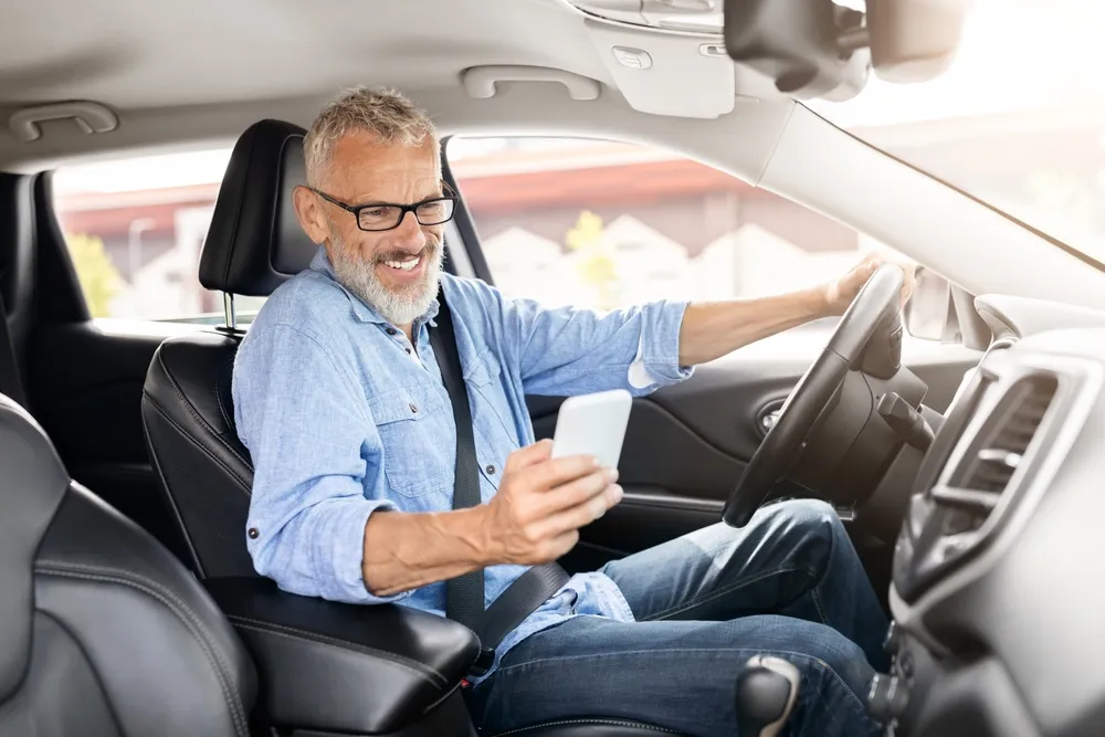 Hombre mayor sonriente, con barba y gafas, sentado al volante de un coche, usando un teléfono móvil.