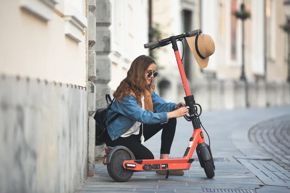 Mujer con chaqueta de mezclilla y gafas de sol ajustando un patinete eléctrico en una calle empedrada. Un sombrero descansa en el manillar.