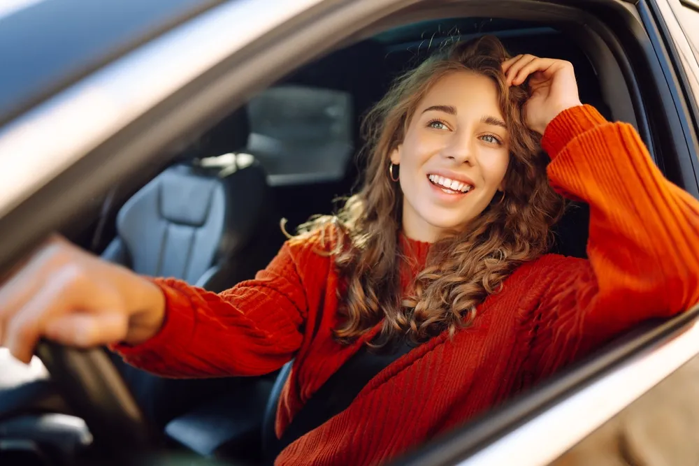 Mujer sonriente con cabello rizado asomándose por la ventana de un coche, vistiendo un suéter rojo.
