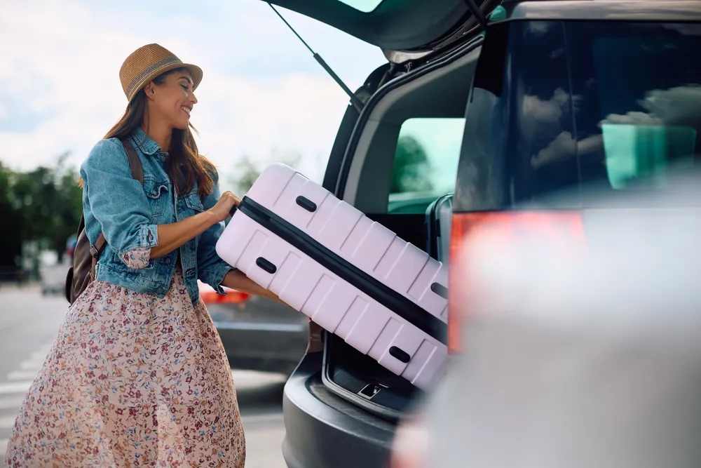 Mujer sonriente con sombrero y vestido floral cargando una maleta rosa en el maletero de un coche.
