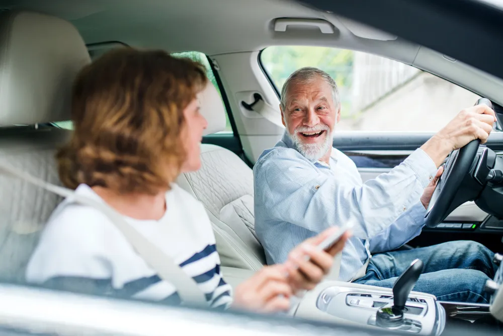 Pareja mayor sonriente en un coche, el hombre conduce mientras la mujer usa su teléfono móvil.