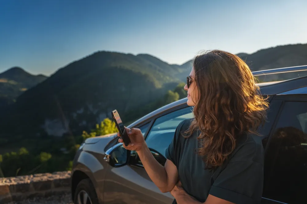 Mujer con cabello rizado y gafas de sol, mirando su teléfono junto a un coche, con montañas al fondo y luz del atardecer.