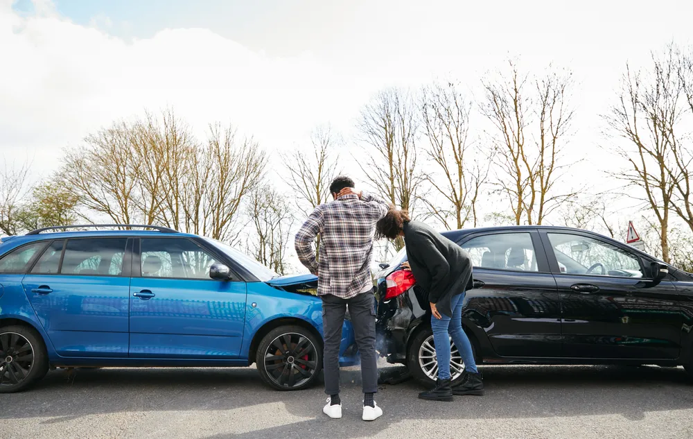 Dos personas observan los daños en sus coches tras un accidente, con árboles al fondo y un cielo parcialmente nublado.