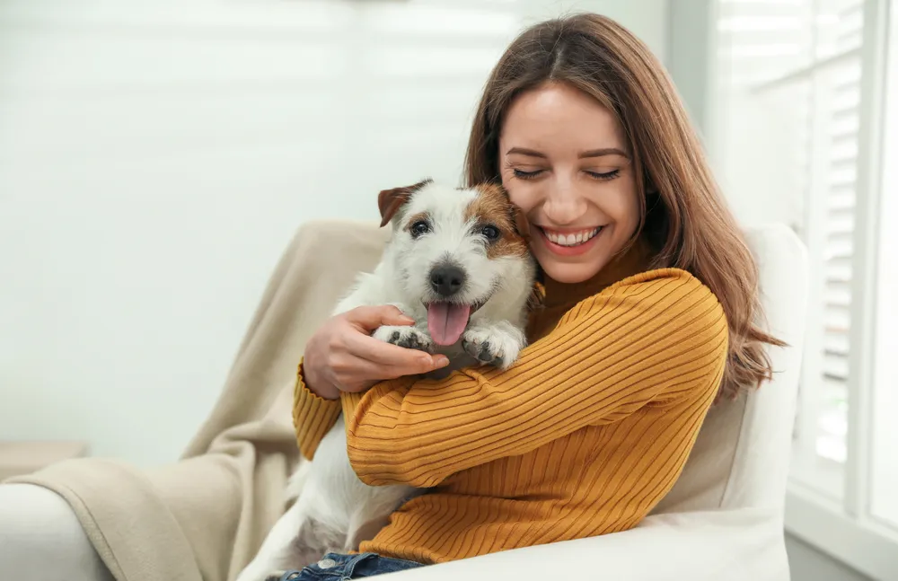 Mujer sonriente abraza a un perro pequeño en un sillón, con luz natural y un ambiente acogedor.