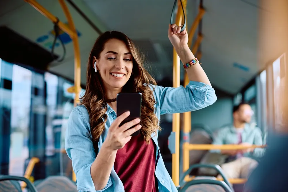 Mujer sonriente en un autobús, usando auriculares y mirando su teléfono móvil.
